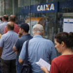 Pessoas em fila na porta de agência da Caixa com aviso na entrada, banco público, sistema financeiro