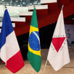 Três bandeiras em ambiente corporativo: França (azul, branco, vermelho), Brasil (verde, amarelo, azul) e Minas Gerais, representando cooperação econômica internacional
