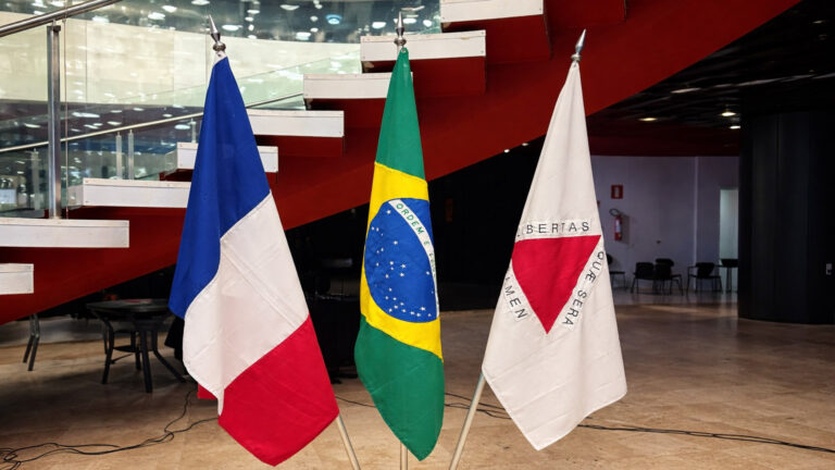 Três bandeiras em ambiente corporativo: França (azul, branco, vermelho), Brasil (verde, amarelo, azul) e Minas Gerais, representando cooperação econômica internacional