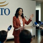 Executiva em terno vermelho durante coletiva de imprensa na sede da OMC com microfones e jornalistas presentes comércio internacional