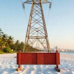 Torre de transmissão de energia elétrica instalada na areia da praia com coqueiros e cidade ao fundo - setor elétrico