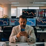 Executivo de terno bege usando smartphone em mesa de operações com monitores exibindo gráficos de bolsa e cotações financeiras