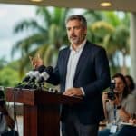 Homem de terno discursa em púlpito cercado por jornalistas e microfones, política econômica governo brasileiro