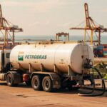 Caminhão-tanque da Petrobras BR em porto com guindastes e contêineres ao fundo, operação petrolífera exportação