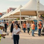 Evento corporativo ao ar livre com profissionais em coletes laranja, tendas brancas e estruturas industriais economia
