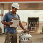 Trabalhador com capacete branco segura botijão de GLP 13kg e prancheta em posto de combustível, subsídio energia