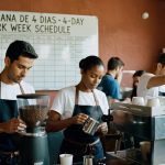 Baristas uniformizados preparam café em estabelecimento com quadro de horários de trabalho ao fundo, mostrando jornada laboral reduzida