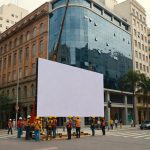 Trabalhadores instalando grande tela branca em cruzamento urbano cercado por edifícios comerciais investimento privado